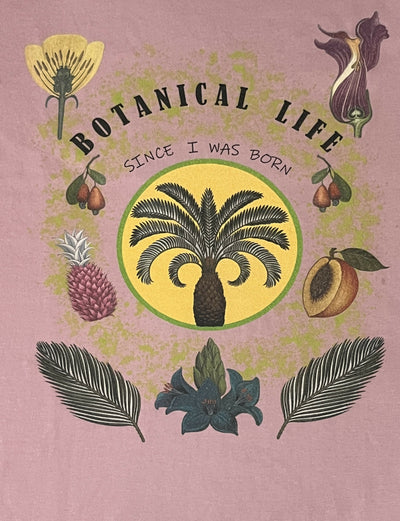 ハラカドのオーガニックコットン ボタニカルライフT＜D.PINK＞は、パープル地に「BOTANICAL LIFE SINCE I WAS BORN」の文字を中心に、ヤシの木や花、松ぼっくり、葉など、さまざまな植物のイラストをグラフィックに落とし込んだデザイン。オーガニックの価値観を体現し、エシカルなライフスタイルを提案するデザインです。