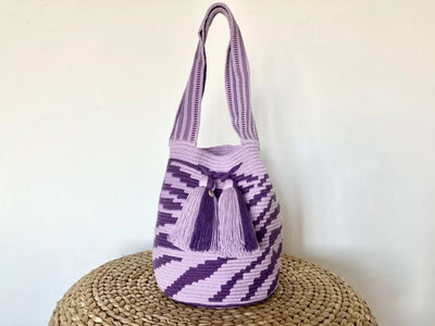 ハラカドのコロンビア発 Pumba Bag Large Purple アクリル 100% ショルダー バッグ。2 つの装飾的なタッセルと斜めのストライプ パターンを備えた手作りのラベンダーと紫のデザインが特徴です。オーガニック素材で作られたこのバッグはプレゼントに最適です。バッグは、無地の白い背景に対して織られたスツールの上に置かれています。