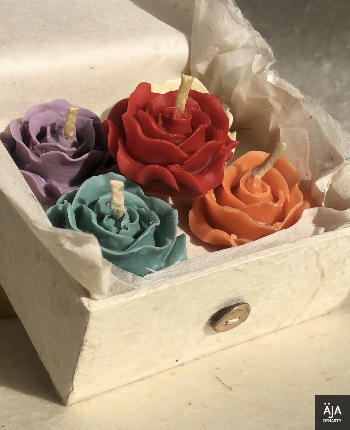 小さな開いた箱には、ハラカドの「Thimi-Tea-Light-Holder 薔薇型のキャンドル 1 点付き」が入っています。紫、赤、オレンジ、ティールの異なる色の 4 つのバラの形のキャンドルが特徴です。これらは丸めたクッキングシートのような紙の上に置かれており、右下隅に「AJA DYNASTY」と書かれたロゴが部分的に見えています。あらゆる機会にサステナブルな贈り物として最適です。