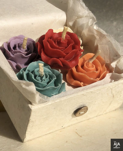 小さな開いた箱には、ハラカドの「Thimi-Tea-Light-Holder 薔薇型のキャンドル 1 点付き」が入っています。紫、赤、オレンジ、ティールの異なる色の 4 つのバラの形のキャンドルが特徴です。これらは丸めたクッキングシートのような紙の上に置かれており、右下隅に「AJA DYNASTY」と書かれたロゴが部分的に見えています。あらゆる機会にサステナブルな贈り物として最適です。