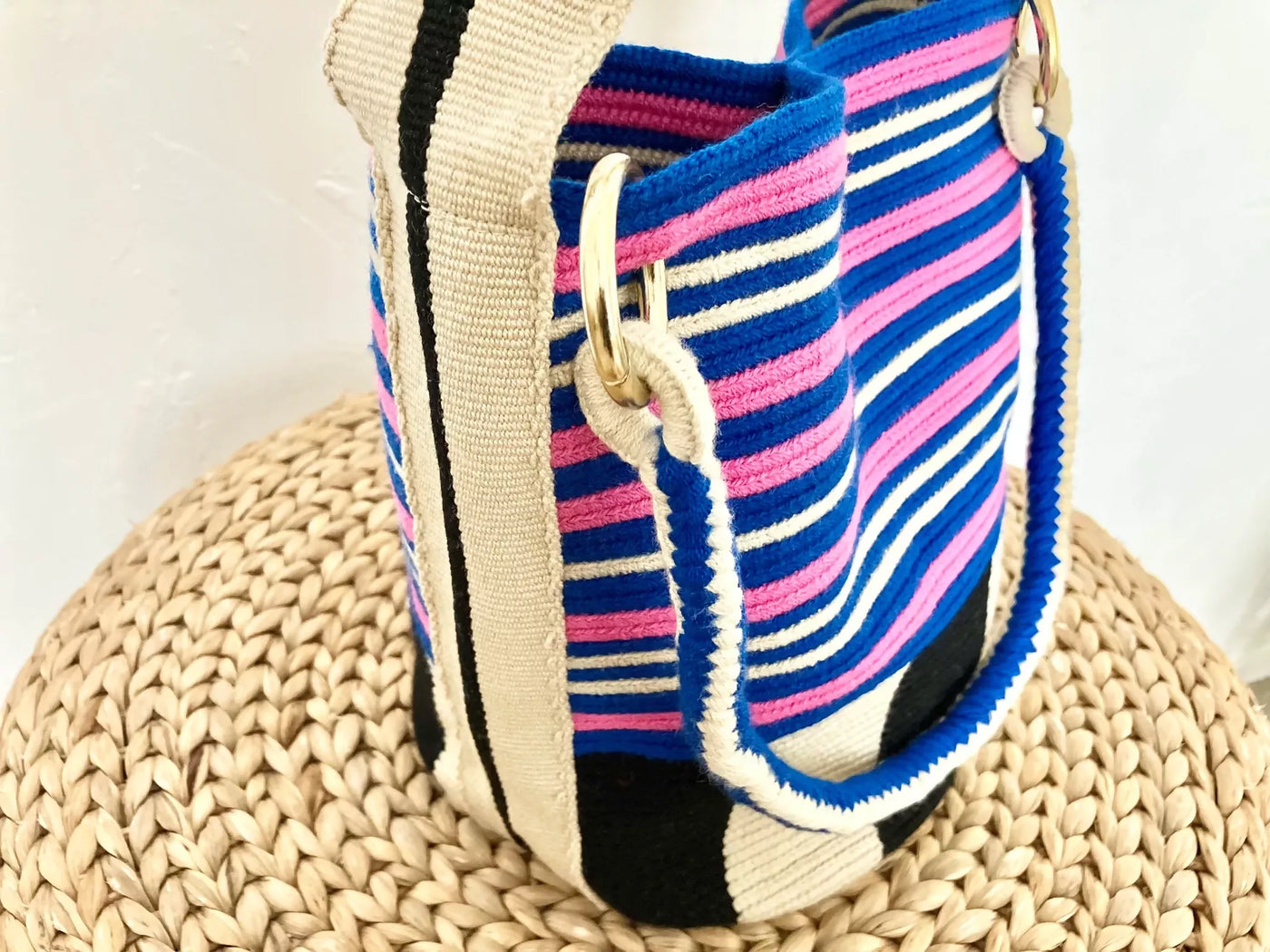 ハラカドのコロンビア発 Arequipe Bag Blue アクリル 100% は、鮮やかな青、ピンク、黒、白の横縞が特徴で、丸いストロー編みの表面に表示されています。サステナブルバッグには、ゴールドのリングに取り付けられた丈夫なベージュのストラップが付いています。